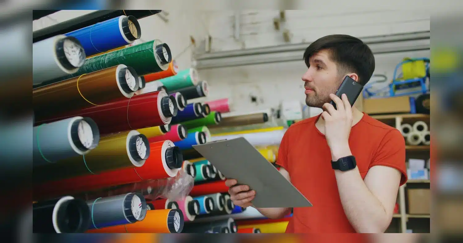 Vinyl Quality Guide — Young man talking on phone, holding a clipboard in a print shop filled with colorful rolls.