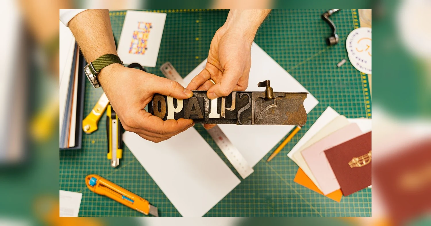 print ready file setup — Hands holding a vintage letterpress tool over a creative workspace with various craft supplies.